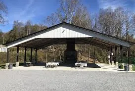 Outdoor pavilion with black roof and central stone fireplace, surrounded by gravel area and trees in the background. No human shapes.