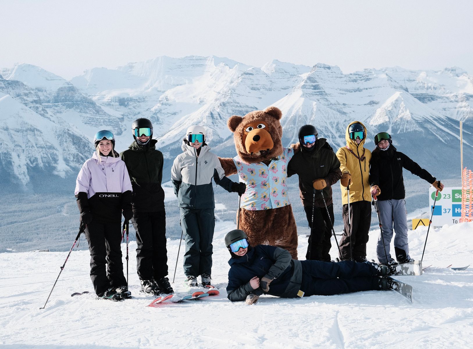 The Bear Street Outfitters team and the Bear Street bear on the slopes, with snowy mountains behind