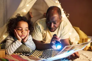 Father and daughter laying in tent reading a book by flashlight.