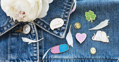 Examples of hard enamel pins on a jean jacket. Pins include a pink and blue pill, word bubble that says "I'm happy", pink heart, four leaf clover, and two cats snuggling against each other. Photo courtesy of Canva.